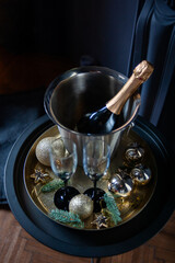 Stylish New Year celebration setup with a champagne bottle in a silver ice bucket, two elegant glasses, gold ornaments, and pine branches on a golden tray, set against a dark, luxurious background