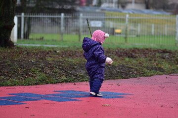 Obraz premium Baby girl standing backwards and holding tree branch in children playground in cold weather autumn winter