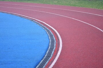 red running track curve with white lines, blue sports field in left side and green grass in right side
