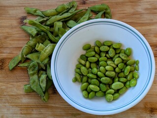 Fresh Shelled Edamame Beans in White Bowl with Empty Pods on Wooden Cutting Board