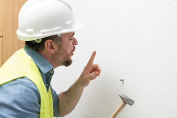 A worker in a white helmet and yellow reflective vest accidentally hits his finger with a hammer while attempting to drive a nail into a wall, illustrating a moment of pain and the risks of manual lab