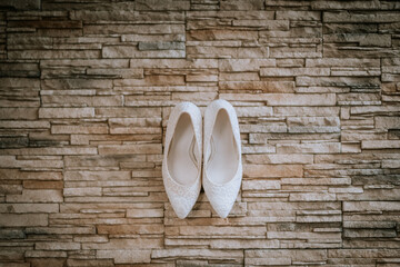 A pair of white lace bridal shoes neatly arranged on a textured stone wall backdrop, creating a stylish and elegant composition.