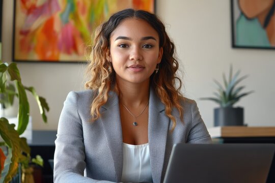 Confident young businesswoman working on laptop in modern office, looking directly at the camera.