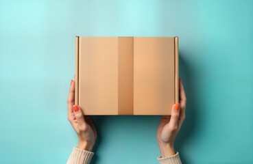 Female hands hold light brown cardboard box. Top view mockup on pastel blue background. Eco-friendly packaging concept for online shopping, delivery. Package likely for gift order. Modern