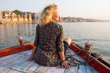 Blonde woman enjoying a boat ride on Ganges river at sunset overlooking the riverfront of Varanasi...
