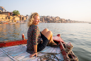 Blonde woman enjoying a boat ride on Ganges river at sunset overlooking the riverfront of Varanasi ancient town in Uttar Pradesh, India