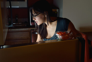 Attractive Hispanic woman looking in refrigerator at night - late night snack - holding cut fruit