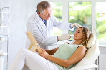 Fototapeta premium Mature man cosmetologist examines middle-aged female patients face after lifting procedure and gently palpates touches skin with finger