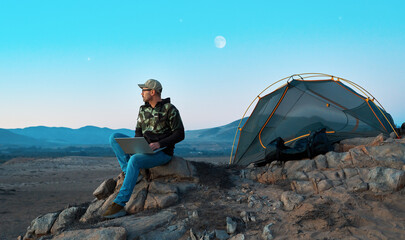Digital nomad working on laptop near his tent in the mountains at night