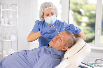 Female doctor in disposable mask inserts filler into patients nasal bridge area for mature male...