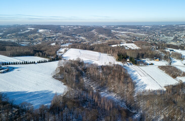 Aerial view of a serene winter landscape with snow-covered hills and bare trees under a clear blue sky.