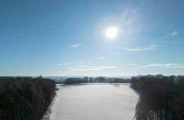 A serene winter landscape featuring a snowy field under a clear blue sky and a bright sun. The...