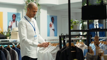 Shopping center employee organizing new fashion collection on racks, ensuring a look book concept for customers seeking trendy apparel in an urban retail space. Modern shopping experience. Camera A.