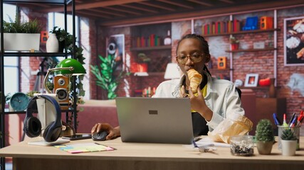 Person enjoying leisure time at home, eating snacks, texting friends on laptop. African american woman enjoying croissant, talking with mates on social network online website, using notebook, camera B