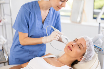 Obraz premium Young adult female patient receiving professional hardware facial procedure at beauty clinic using cooling attachment to alleviate inflammation, rejuvenate of skin and enhance complexion