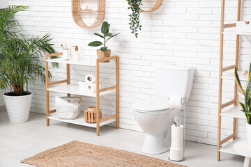 Interior of restroom with toilet bowl, shelving units and houseplants near white brick wall