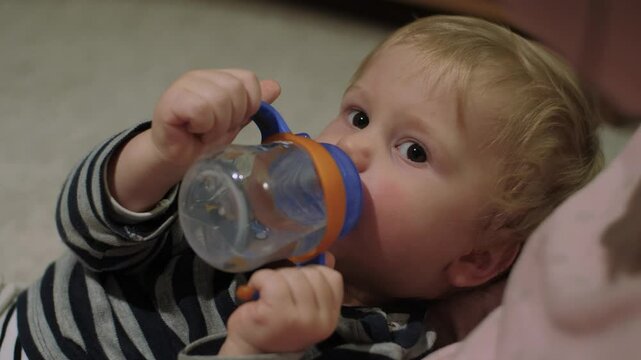 Cute and innocent toddler lies comfortably and joyfully sipping from colorful sippy cup. Child looking at camera showcasing pure joy of childhood and essential importance of staying hydrated