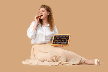 Beautiful young woman with box of sweet chocolate candies sitting on beige background
