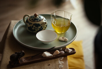 Still life photo of patterned clay teapot, clear glass pitcher, tea cup with fresh brewed oolong tea on tray in ceremony room. Soft light, wooden tea tray evoke calming mindful Chenese atmosphere.