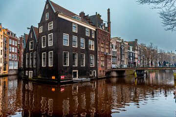 Historic Dutch canal house with black facade reflecting in the water of an Amsterdam canal. A picturesque bridge and colorful neighboring buildings