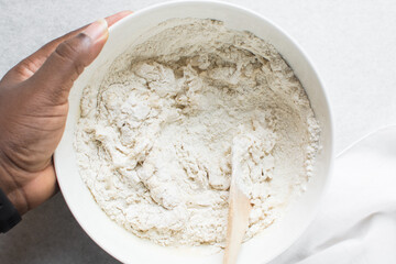 Overhead view of bread dough being mixed in a white bowl, top view of homemade bread dough being mixed on white granite countertop, process of making artisan bread
