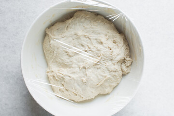 Overhead view of bread dough in a whiite mixing bowl covered with plastic wrap, dough rising in a ceramic bowl, process of making artisan bread