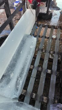 Man cuts a clear, rectangular block of ice with an icepick on a metal trailer, creating a spray of ice particles