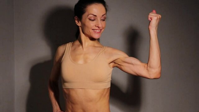 Healthy smiling woman posing and showing strong strength biceps muscle, attractive body. Slim caucasian female model posing in beige bra in studio low key light on wall background. Sporty lifestyle