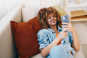 Holding Phone, Smiling Woman, Relaxing on Cozy Sofa in Home: Indoor Portrait of Beautiful Young...