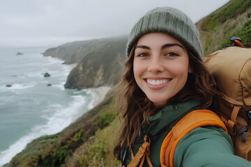 A selfie of an attractive brunette woman hiking on the coast, wearing green and orange outdoor gear with a brown backpack, smiling at the camera