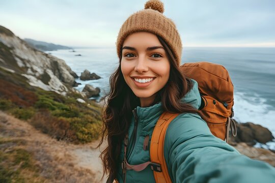 A selfie of an attractive brunette woman hiking on the coast, wearing green and orange outdoor gear with a brown backpack, smiling at the camera - Powered by Adobe