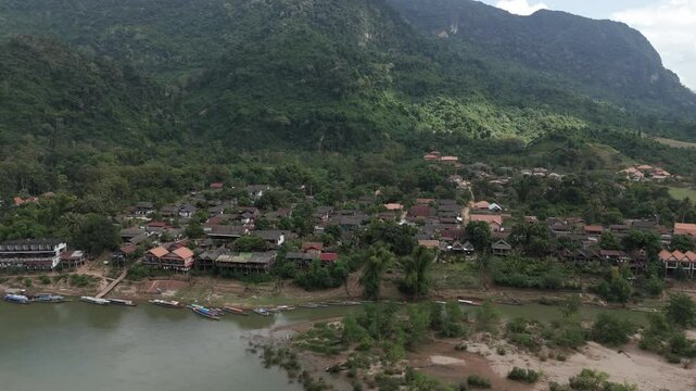 Spinning drone footage in front of a traditional wooden village of north Laos, Muang Ngoy. Camera is moving from left to right showing river and mountains in the background.