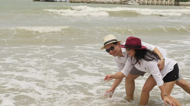 A couple wearing hats, enjoying playful moments at the beach, bending near the shoreline while interacting with the gentle waves on a bright, breezy day