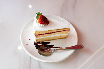 Slice of layered cake with white frosting and a strawberry on top sits on a white plate. A fork rests beside it. The cake appears to have a fruit filling, possibly strawberry matching the garnish.