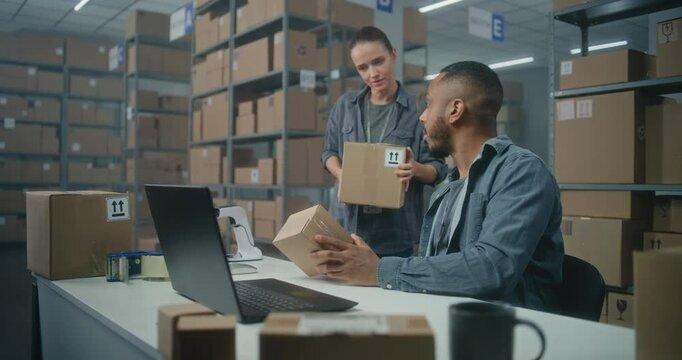 African American logistics manager scans parcel with barcode scanner, talks to colleague, uses laptop. Sorting center employee carrying boxes for shipping. Storage of E-commerce store or post office.