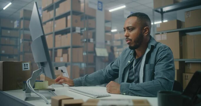 African American logistics postal service employee checks online orders, works on computer. Sorting center female worker checking parcels for delivery to customers. Warehouse or distribution center.