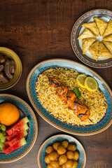Iftar food consists of Chicken biryani, fruits and Mediterranean dessert. Arabic dishes are arranged on the dark wood background, shot from top angle. 