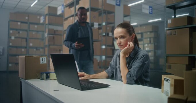 Warehouse or sorting center: Female logistics specialist checks E-commerce online orders on laptop. African American warehouse associate uses tablet computer, consults with colleague. Delivery service
