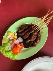 Goat and deer meat satay seasoned with rich spices, then grilled to perfection and served with sliced ​​cabbage, tomatoes, shallots, cayenne pepper, and lime to add to the flavor on green plate.