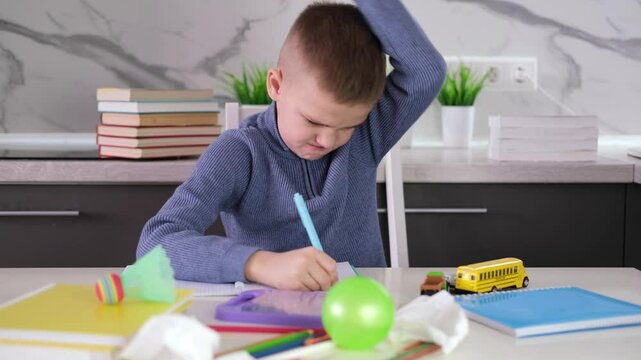  Frustrated, unhappy child sits down at table and does his homework among stack of books and scattered pieces of paper and toys. Education school, learning disabilities, reading difficulties, dyslexia