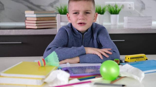 Frustrated, unhappy child sits down at table and does his homework among stack of books and scattered pieces of paper and toys. Education school, learning disabilities, reading difficulties, dyslexia.