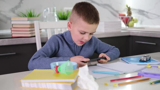  Education, school, learning difficulties, reading difficulties. Child plays with smartphone instead of doing homework. Schoolchild sits at table doing homework among pile of books and scattered toys.