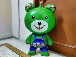 Green bear doll with colorful balloons in front of the wooden door.
