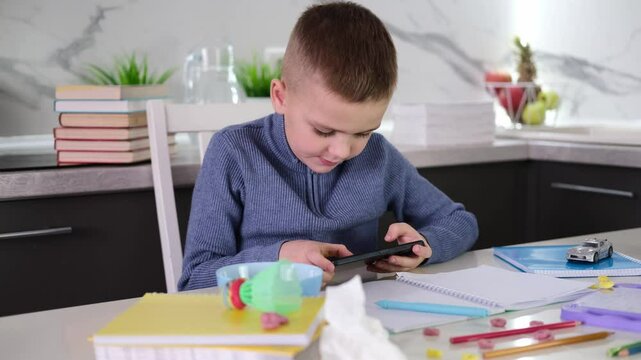 Education, school, learning difficulties, reading difficulties. Child plays with smartphone instead of doing homework. Schoolchild sits at table doing homework among pile of books and scattered toys.