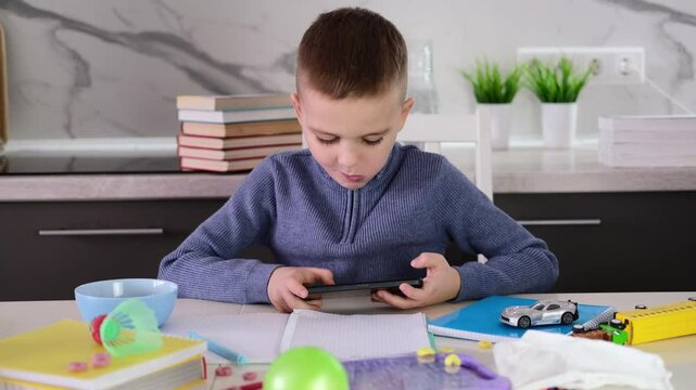 Education, school, learning difficulties, reading difficulties. Child plays with smartphone instead of doing homework. Schoolchild sits at table doing homework among pile of books and scattered toys.