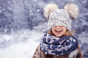 Winter Children Fun Holidays. Happy Child Portrait in Winter Nature Background. Emotional Smiling Little Girl Kid in Funny Knitted Hat on Snow Falling Background. Copy Space.