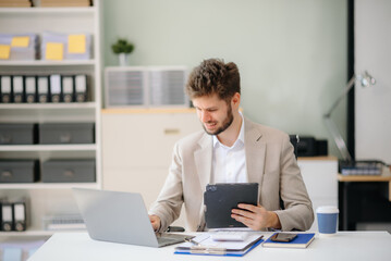 Obraz premium Confident businessman working on finance analysis with a tablet and laptop at office desk.