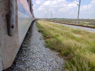 A moving train glides through a scenic landscape. Gravel tracks, grassy embankments, and electric poles stretch into the horizon under a blue, cloud-speckled sky, evoking motion and exploration.