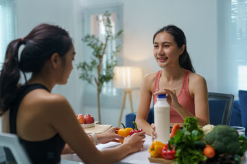 Two sporty young women are sitting at a table, discussing nutrition and a healthy lifestyle while examining fresh vegetables and a bottle of milk