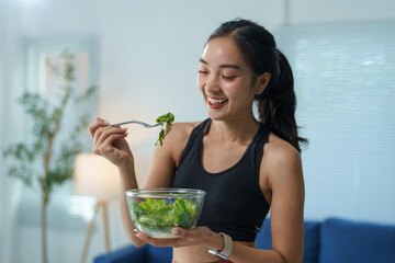 Young sporty asian woman in sportswear is smiling and eating healthy green salad after workout at...